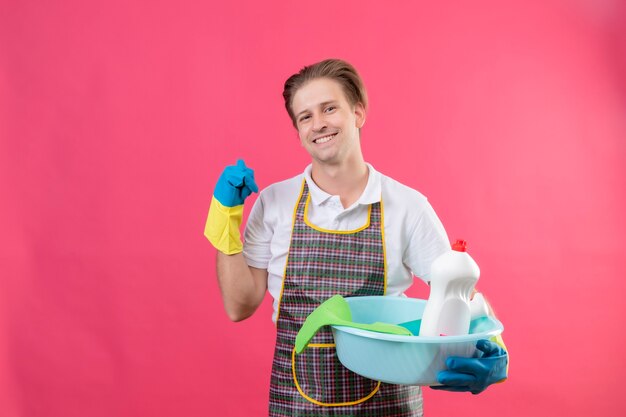 Young hansdome man wearing apron and rubber gloves holding basin with cleaning tools smiling pointing to something behind with thumb smiling standing over pink wall