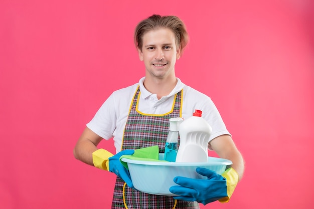 Young hansdome man wearing apron and rubber gloves holding basin with cleaning tools with confident smile on face standing over pink wall