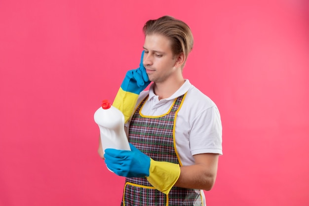 Young hansdome man wearing apron and rubber gloves holding bottle with cleaning supplies looking at it with pensive expression thinking standing over pink wall