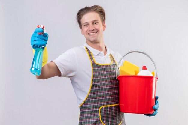 Young hansdome man wearing apron and rubber gloves holding bucket with cleaning tools and cleaning spray