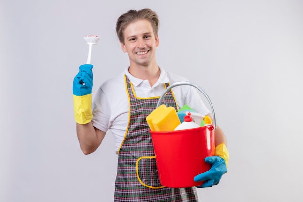 Young hansdome man wearing apron and rubber gloves holding bucket with cleaning tools and scrubbing