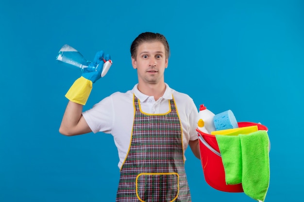 Free photo young hansdome man wearing apron and rubber gloves holding bucket with cleaning tools