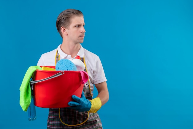 Young hansdome man wearing apron and rubber gloves holding bucket with cleaning tools