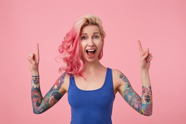 Foto gratuita giovane bella donna dai capelli rosa stupita felice in maglietta blu con le braccia alzate, sente notizie incredibili, con la bocca spalancata per la sorpresa, si alza e indica lo spazio della copia.