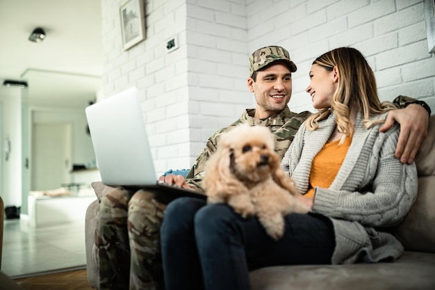 Young happy army soldier and his wife talking to each other while using computer at home.