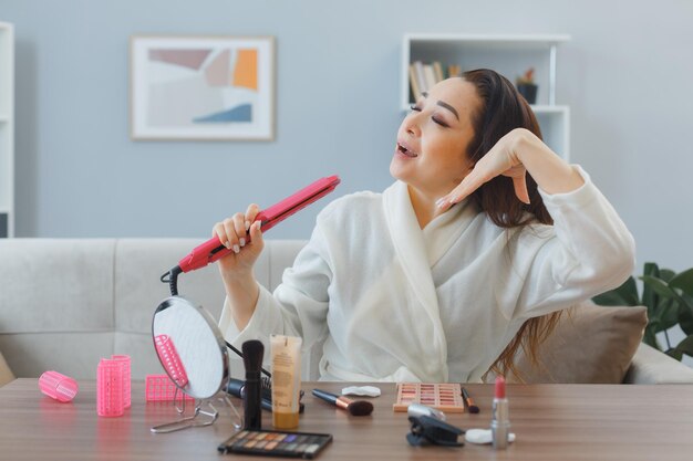 Young happy asian woman with towel in bathrobe sitting at the dressing table at home interior curling iron making styling doing morning makeup routine