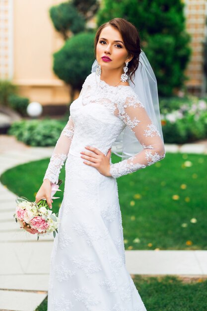 Young happy beautiful bride in white elegant wedding dress with bouquet posing outdoor in park.