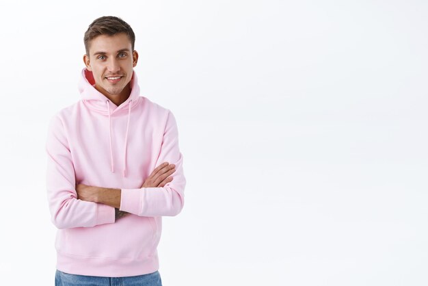 Young happy blond handsome man staying at home in cozy pink hoodie smiling upbeat cross arms chest confident standing white background recommend product on blank copy space
