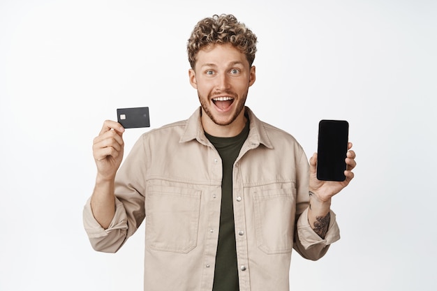 Young happy blond man showing credit card with mobile phone screen smiling amazed announcing awesome promotion standing over white background