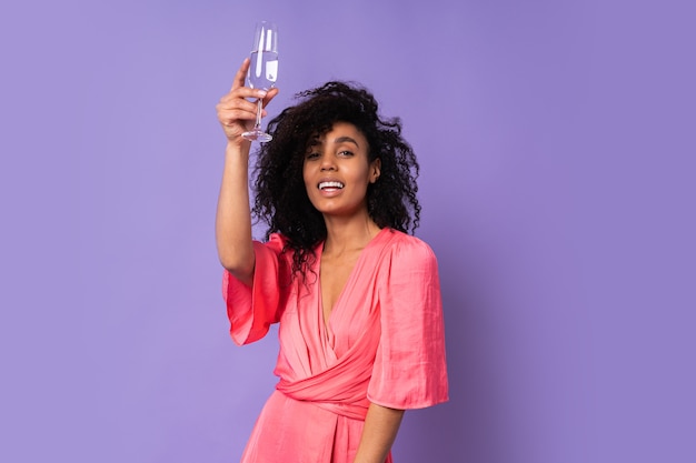 Free photo young  happy  brazillian  female with  curly  hair in  pink stylish dress posing with glass  of 
champagne  over purple wall.  party mood.