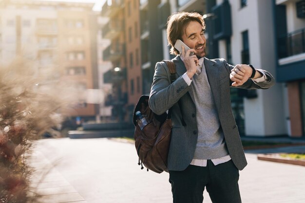 Young happy businessman checking the time on wristwatch while talking on mobile phone in the city