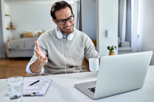 Young happy businessman drinking coffee and using computer while having video conference at home