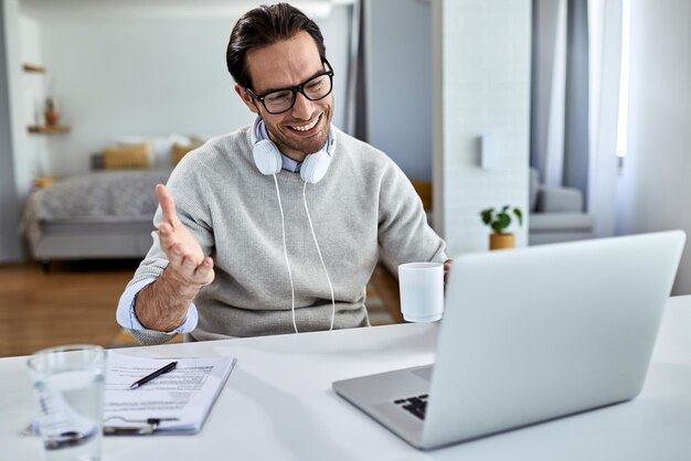 Young happy businessman drinking coffee and using computer while having video conference at home