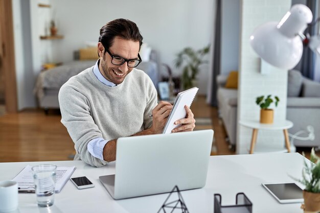 Young happy businessman using computer and writing notes while working at home