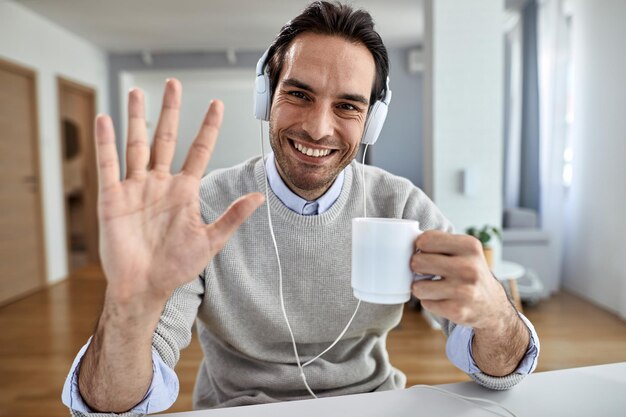 Young happy businessman with headphones waving toward the camera while drinking coffee at home