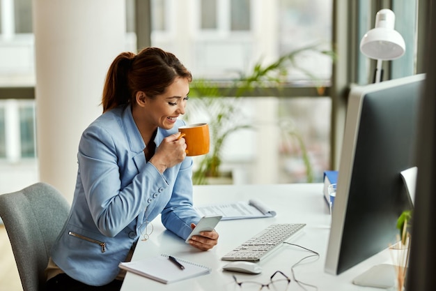 Молодая счастливая деловая женщина пьет кофе и обменивается текстовыми сообщениями по мобильному телефону в офисе
