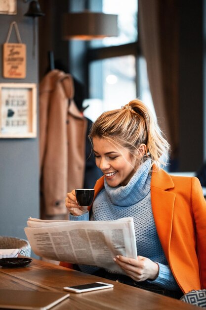 Young happy businesswoman reading daily newspaper and drinking coffee after work in a cafe