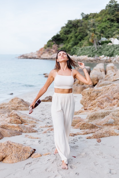 young happy calm caucasian woman with ukulele on tropical rocky beach at sunset