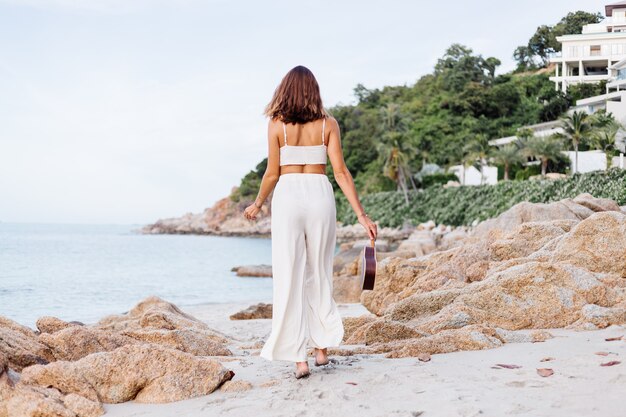 young happy calm caucasian woman with ukulele on tropical rocky beach at sunset