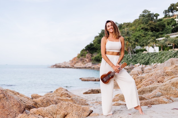 young happy calm caucasian woman with ukulele on tropical rocky beach at sunset