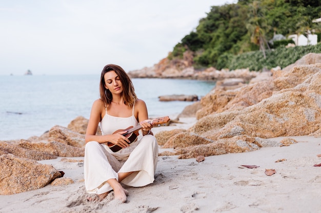 日没時の熱帯の岩のビーチでウクレレと若い幸せな穏やかな白人女性