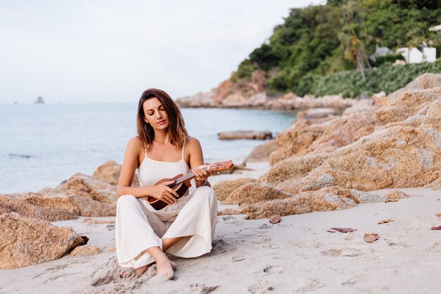 young happy calm caucasian woman with ukulele on tropical rocky beach at sunset