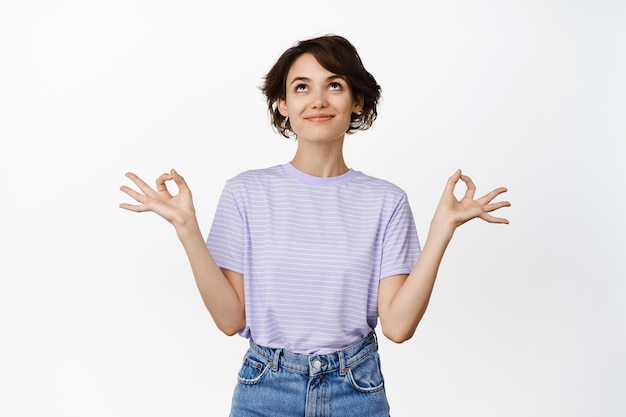 Young happy, calm woman meditating, looking up with relaxed face, holding hands sideways in zen, nirvana lotus pose, practice yoga, standing over white background