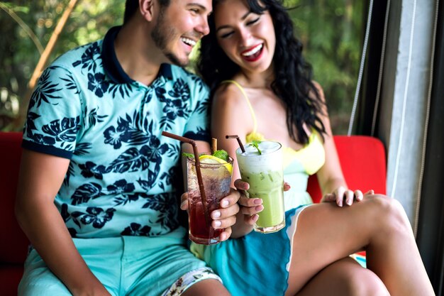 Young happy couple drinking tasty sweet cocktails at tropical bar, smiling and having fun, bright clothes and positive emotions.