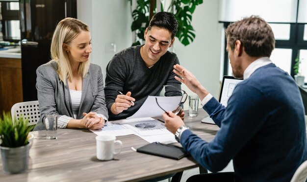 Young happy couple reading terms of mortgage documents while having meeting with real estate agent at home Focus is on young man