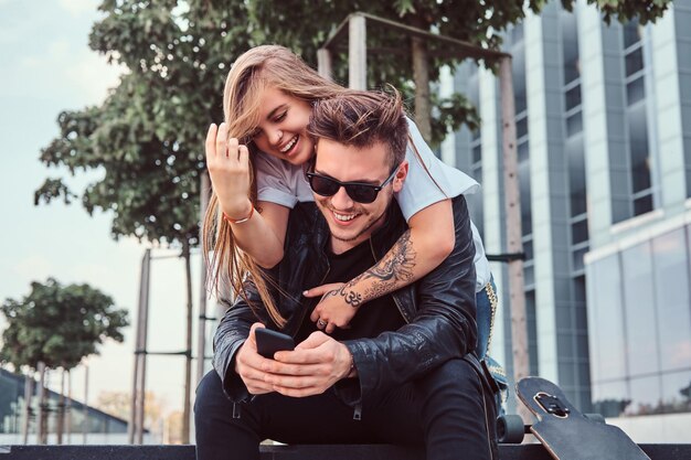 Молодая счастливая пара отдыхает вместе возле небоскреба - красивая девушка обнимает своего парня, пока он пользуется смартфоном.