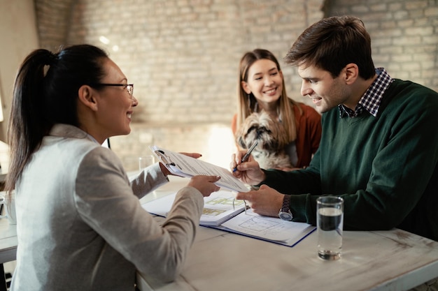 Young happy couple signing document while having meeting with real estate agent in the office Focus is on man