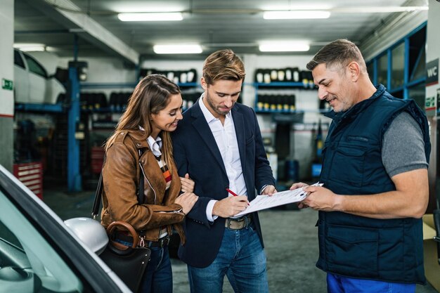 Young happy couple signing documents while being with car mechanic in a repair shop