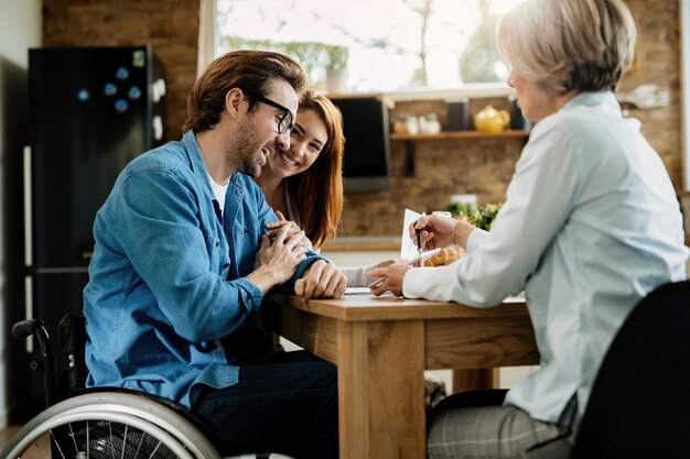Молодая счастливая пара и их финансовый консультант с помощью сенсорной панели на встрече дома В центре внимания мужчина в инвалидной коляске
