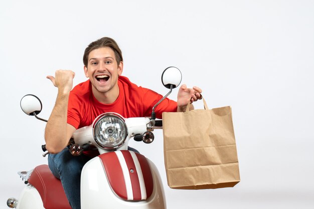 Young happy courier guy in red uniform sitting on scooter holding orders and pointing back on yellow wall