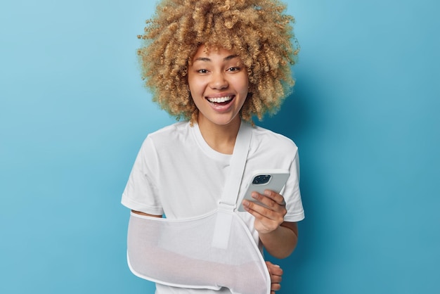 Young happy curly young woman being on sick leave as has broken arm wears sling being injured during dangerous bike riding uses cellphone for chatting with friends poses against blue background