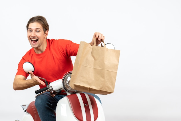 Young happy delivery guy in red uniform sitting on scooter holding paper bag on white wall