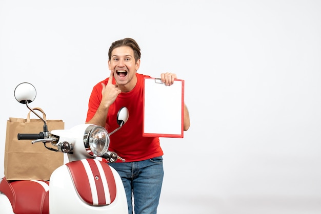 Young happy delivery guy in red uniform standing near scooter holding document on white wall