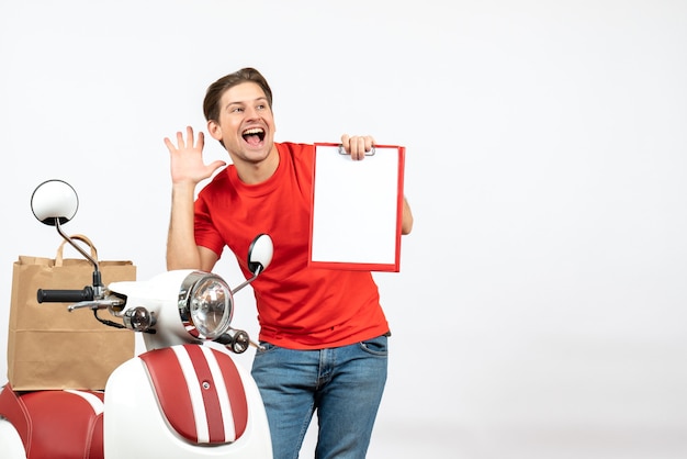 Free photo young happy delivery guy in red uniform standing near scooter showing document on yellow white wall