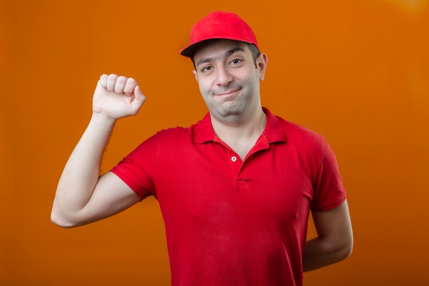 Young happy delivery man in red polo shirt and cap looking confident raising fist after a victory winner concept standing over isolated orange background