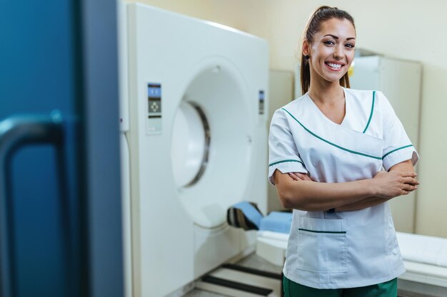 Young happy doctor standing with her arms crossed next to MRI scanner and looking at camera