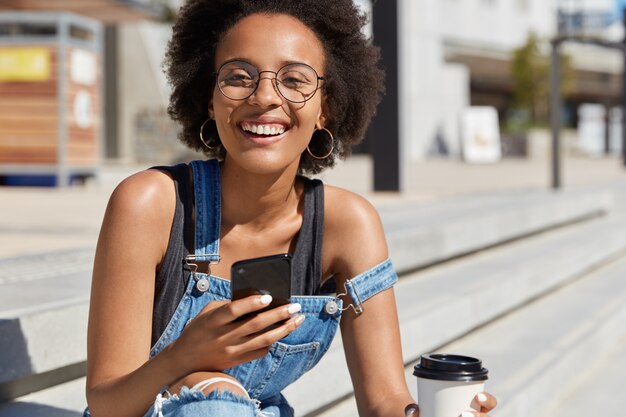 Young happy ethnic female checks email with notification, smiles broadly, poses in urban setting, drinks takeaway coffee, chats on mobile phone, enjoys hot drink. Youth, spare time, technologies