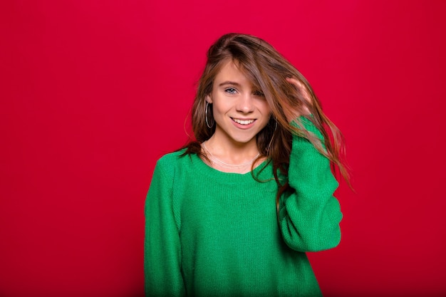 Young happy exited stylish woman with long hair and big eyes wearing green sweater posing with smile on red wall