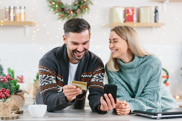 Молодая счастливая семья во время новогодних и рождественских праздников дома радостно покупает подарки онлайн с помощью смартфона и кредитной карты