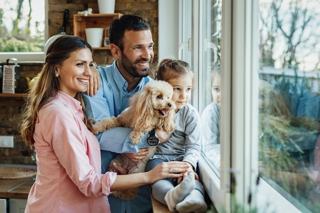 Young happy family and their dg relaxing by the window at home and looking through it