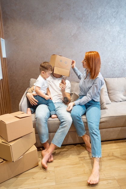 Young happy family with kid unpacking boxes together sitting on sofa