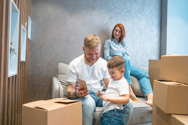 young happy family with kid unpacking boxes together sitting on sofa