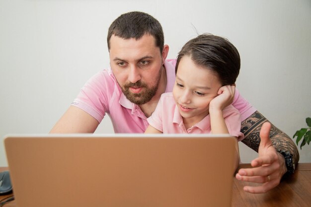 Бесплатное фото Молодой счастливый отец в розовой рубашке поло работает на ноутбуке, сидя за столом со своим маленьким умным сыном счастливая семья работает дома концепция