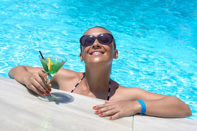 Молодая счастливая женщина в бассейне с бокалом зеленого вкусного коктейля под солнечным светом