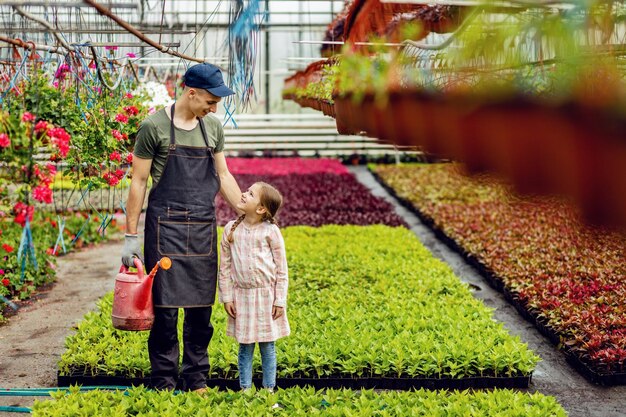 じょうろを持つ若い幸せな花屋は、植物の保育園で小さな女の子と話すことができます