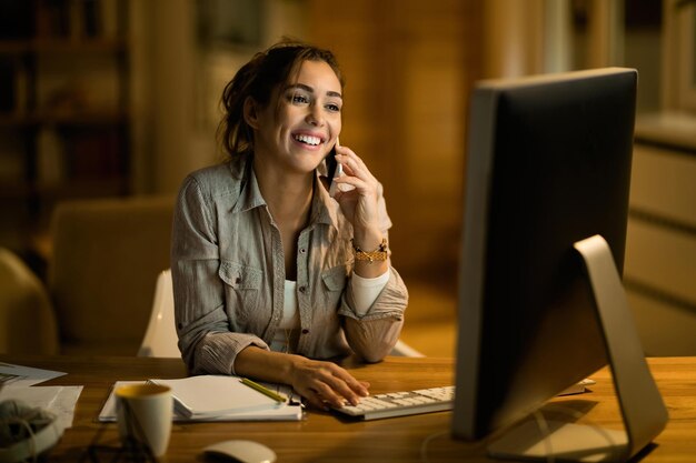 Young happy freelancer communicating on mobile phone while surfing the net on desktop PC at night at home